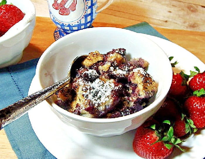 A small white bowl filled with Blueberry Grunt along with a spoon and some fresh strawberries.