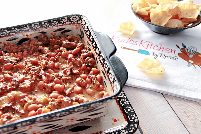 An off set horizontal photo of Cheesy Sausage Bean Dip on the left and a bowl of nacho chips and a logo napkin on the right.