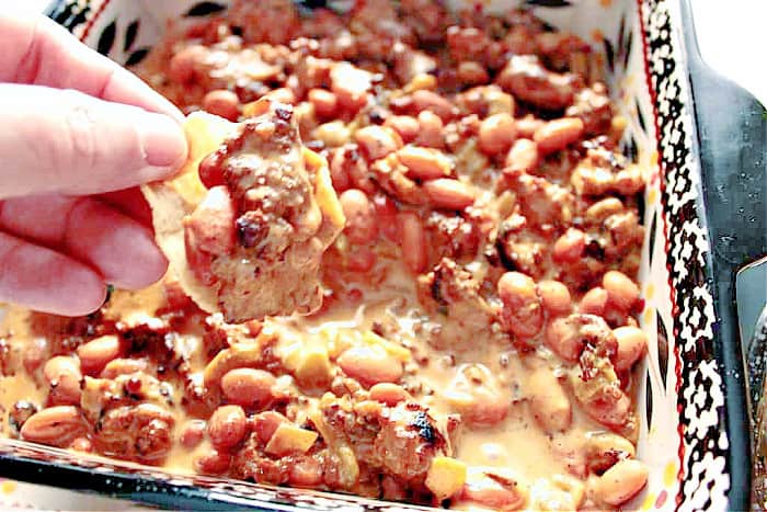 A closeup horizontal photo of a hand holding a chip that's been dipped into sausage bean dip.