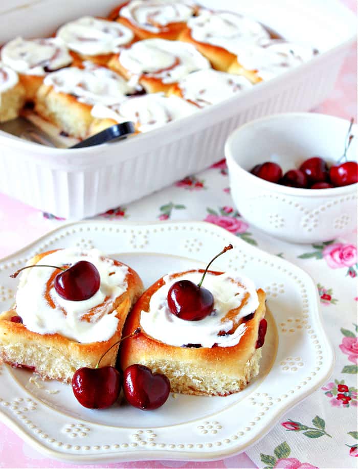 A vertical closeup photo of two Cherry Sweet Rolls on a white plate with cream cheese almond glaze and fresh cherries as garnish.