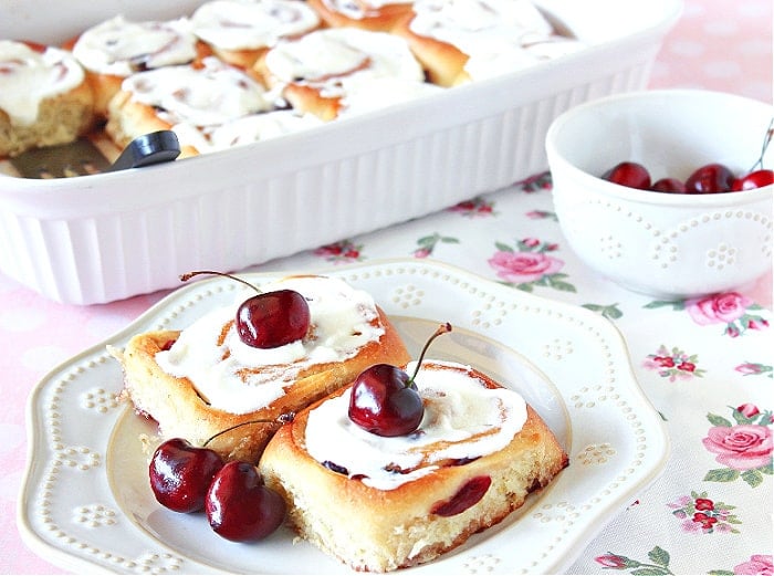 Two Cherry Sweet Rolls on a white plate with a rose bud tablecloth and fresh cherries as garnish on the buns.