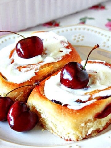 Two iced Cherry Sweet Rolls on a plate with fresh cherries.