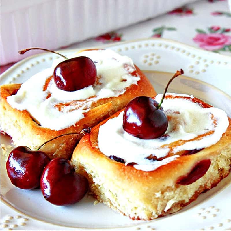 A square closeup photo of two Cherry Breakfast Buns on a white plate with fresh cherries as garnish.