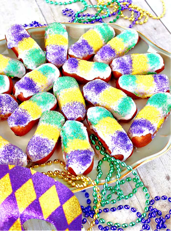 A vertical closeup image of a plate of Mini Long John with colorful sanding sugar and Mardi Gras beads.