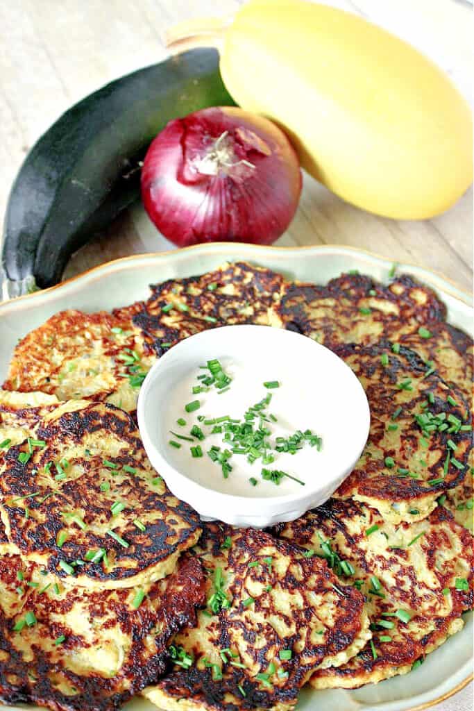 A vertical closeup of a plate of Spaghetti Squash and Zucchini Pancakes with a whole onion, zucchini, and spaghetti squash in the background.