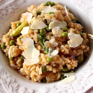 A white bowl filled with Asparagus and Pea Risotto along with shards of Parmesan cheese on top.