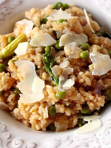A white bowl filled with Asparagus and Pea Risotto along with shards of Parmesan cheese on top.