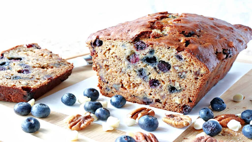 A sliced loaf of Blueberry Banana Quick Bread along with fresh blueberries, pecans, and white chocolate chips on the table.