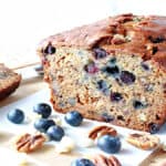 A sliced loaf of Blueberry Banana Quick Bread along with fresh blueberries, pecans, and white chocolate chips on the table.