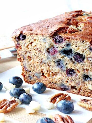 A sliced loaf of Blueberry Banana Quick Bread along with fresh blueberries, pecans, and white chocolate chips on the table.