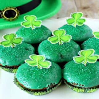 A white scalloped plate filled with Chocolate Mint Brownie Cupcakes with a green glaze and paper shamrocks on top.