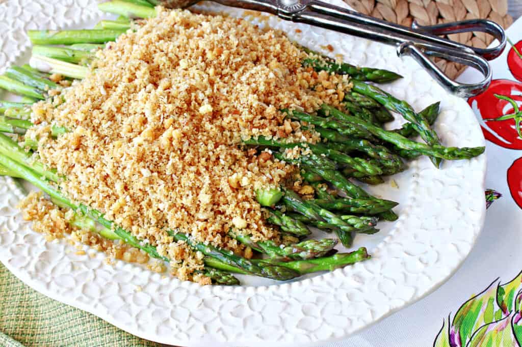 An offset horizontal photo of Asparagus with Breadcrumbs on a pretty white platter with a set of serving tongs on the side of the platter.