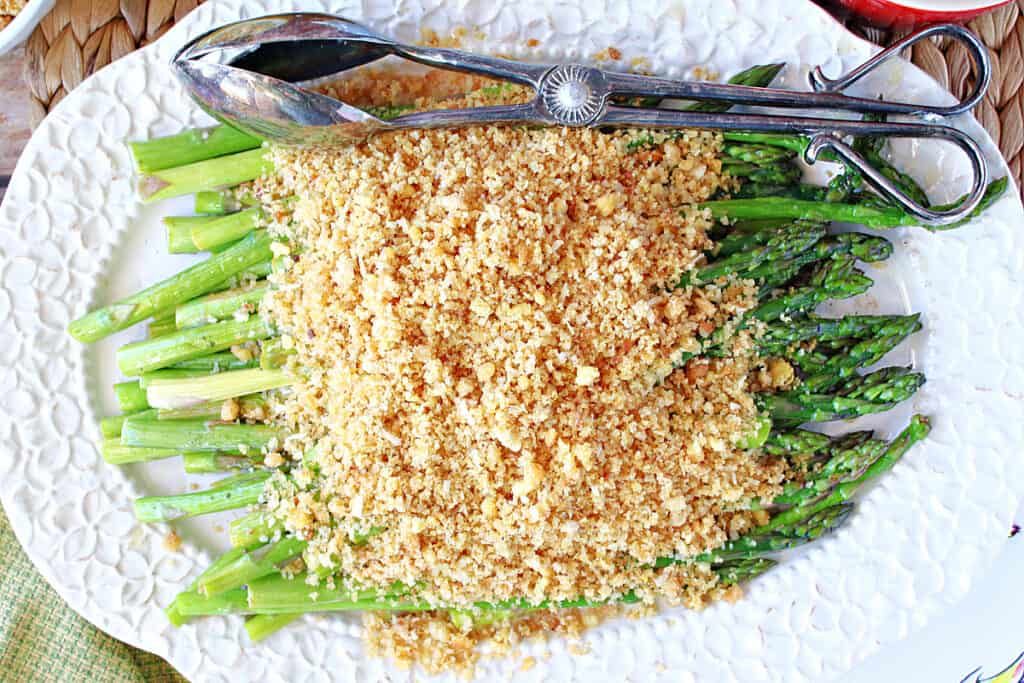 A directly overhead photo of a pretty white platter filled with Asparagus with Breadcrumbs along with a serving tong on the side of the platter.