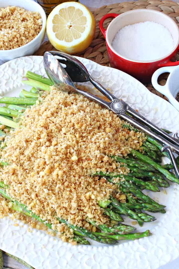 A overhead vertical closeup photo of Asparagus with Breadcrumbs on a pretty white platter with serving tongs.