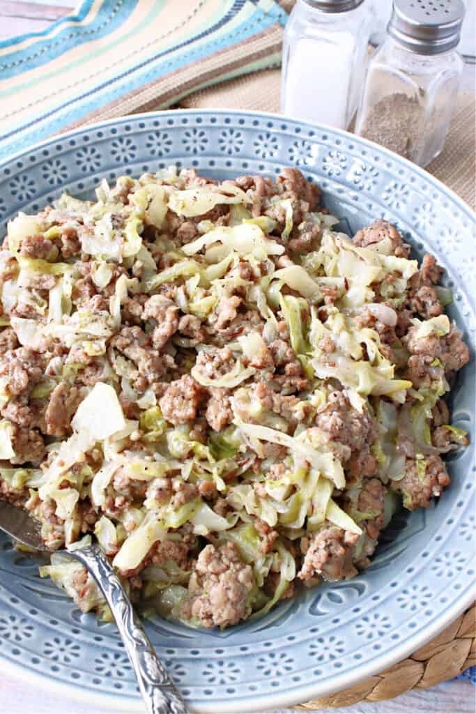A vertical closeup of Ground Pork and Cabbage Skillet in a blue bowl with a spoon and a salt and pepper shaker in the background.
