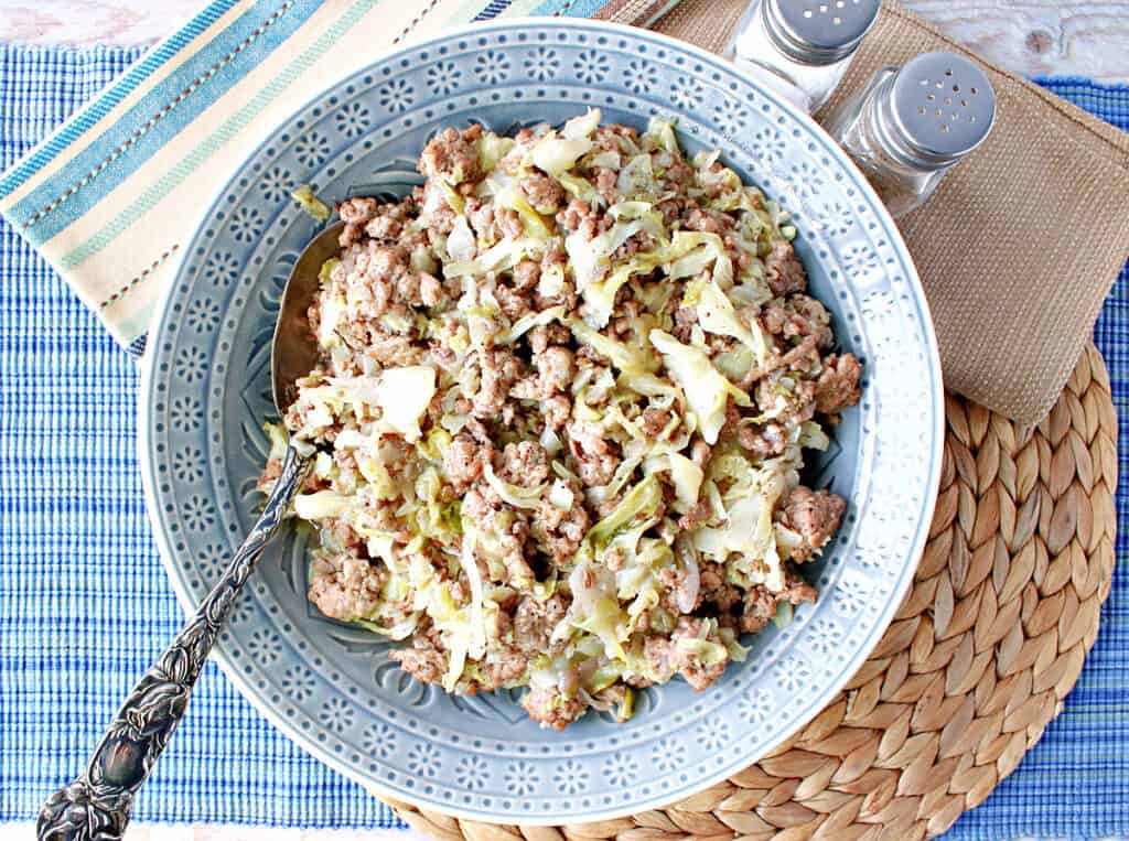 Direct overhead photo of a blue bowl filled with Ground Pork and Cabbage Skillet along with blue and brown napkins and salt and pepper shakers.