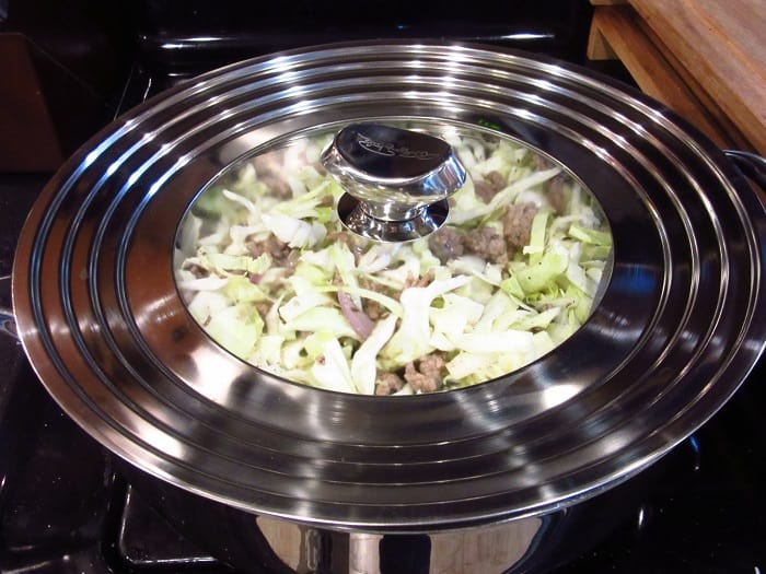 A lid on a large skillet with pork and cabbage.