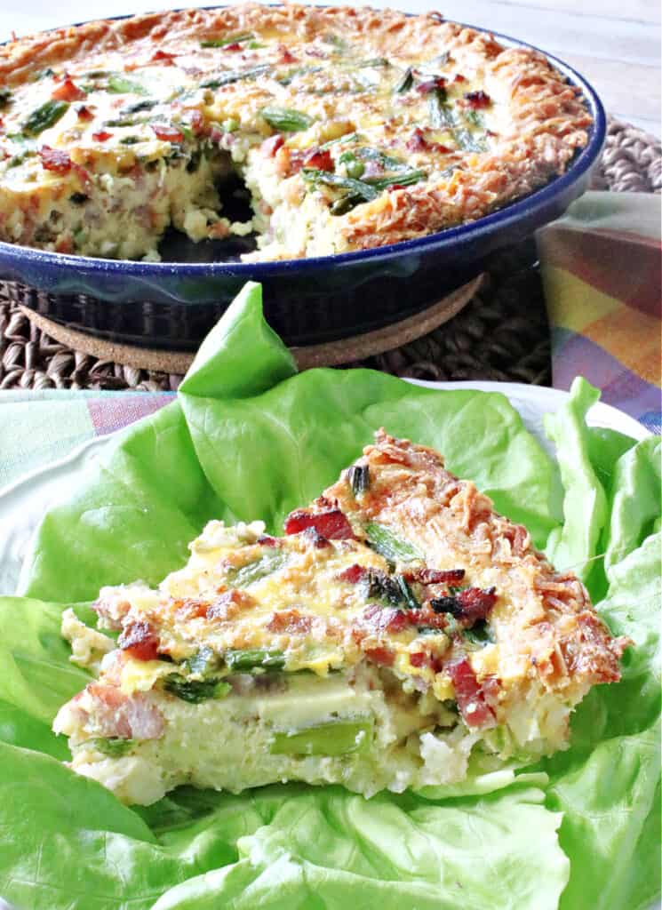 A vertical closeup of a slice of Ham and Asparagus Quiche in the foreground on top of lettuce and a whole quiche in the background.