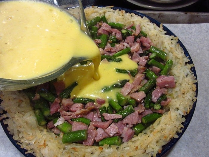 Eggs being poured into a ham and asparagus quiche with a hash brown crust.