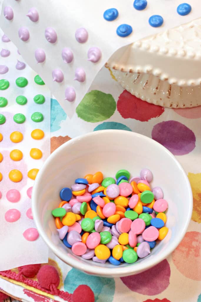 A vertical closeup of colorful homemade candy buttons in a bowl and also on paper strips in the background.