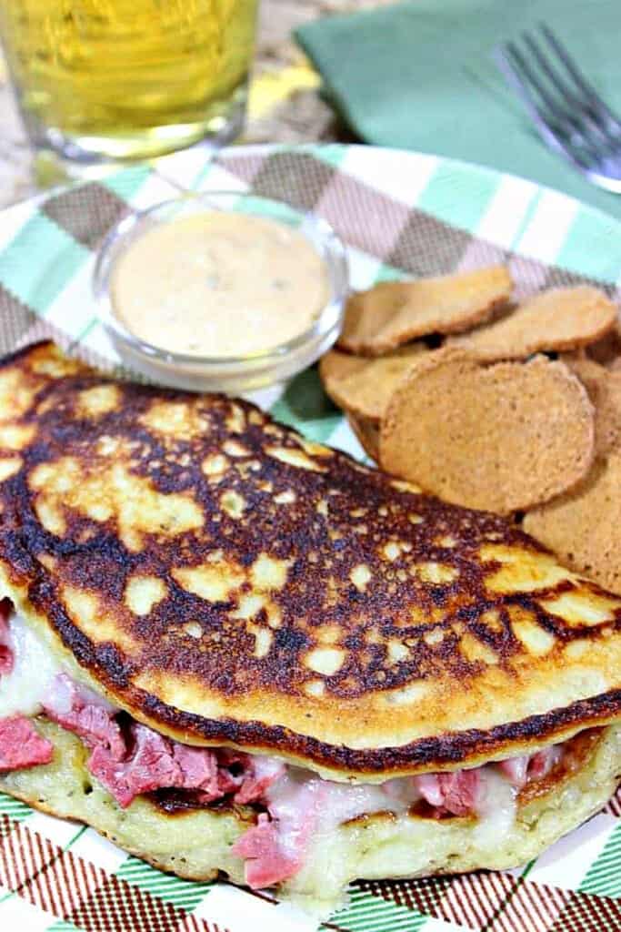 A vertical closeup of and Irish potato pancake filled with corned beef and Swiss cheese along with chips and a beer in the background.