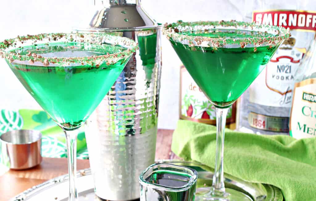 Two right green Irish Grasshopper Cocktails in martini glasses along with a shot glass filled with crème de menthe and a cocktail shaker in the background.