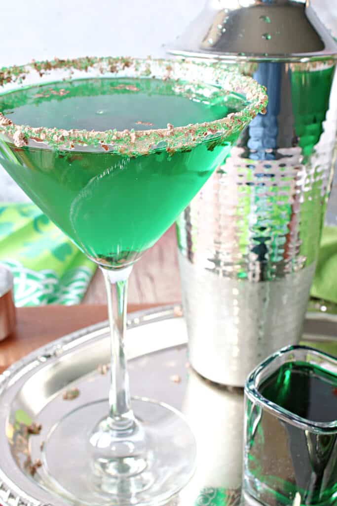 A vertical closeup photo of a martini glass filled with a green Irish Grasshopper Cocktail, and a cocktail shaker on a silver platter.