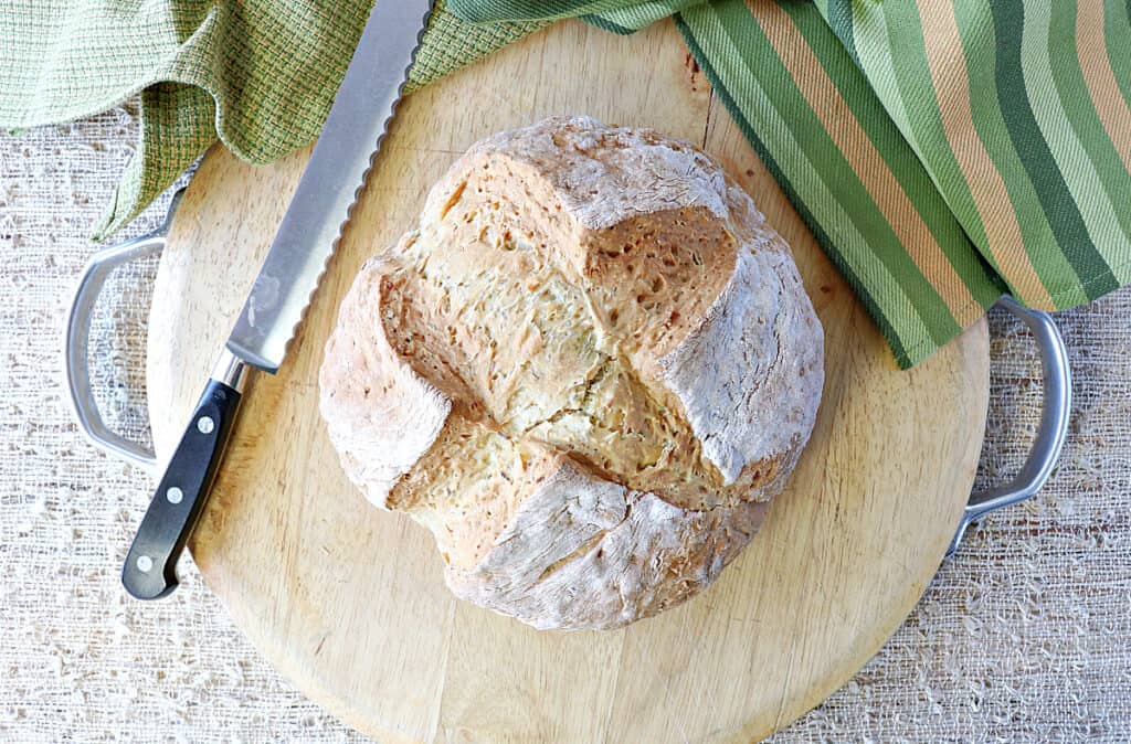 A direct overhead photo of a loaf of Irish Soda Bread with Caraway and Dill on a wooden cutting board with a serrated knife and some green napkins.