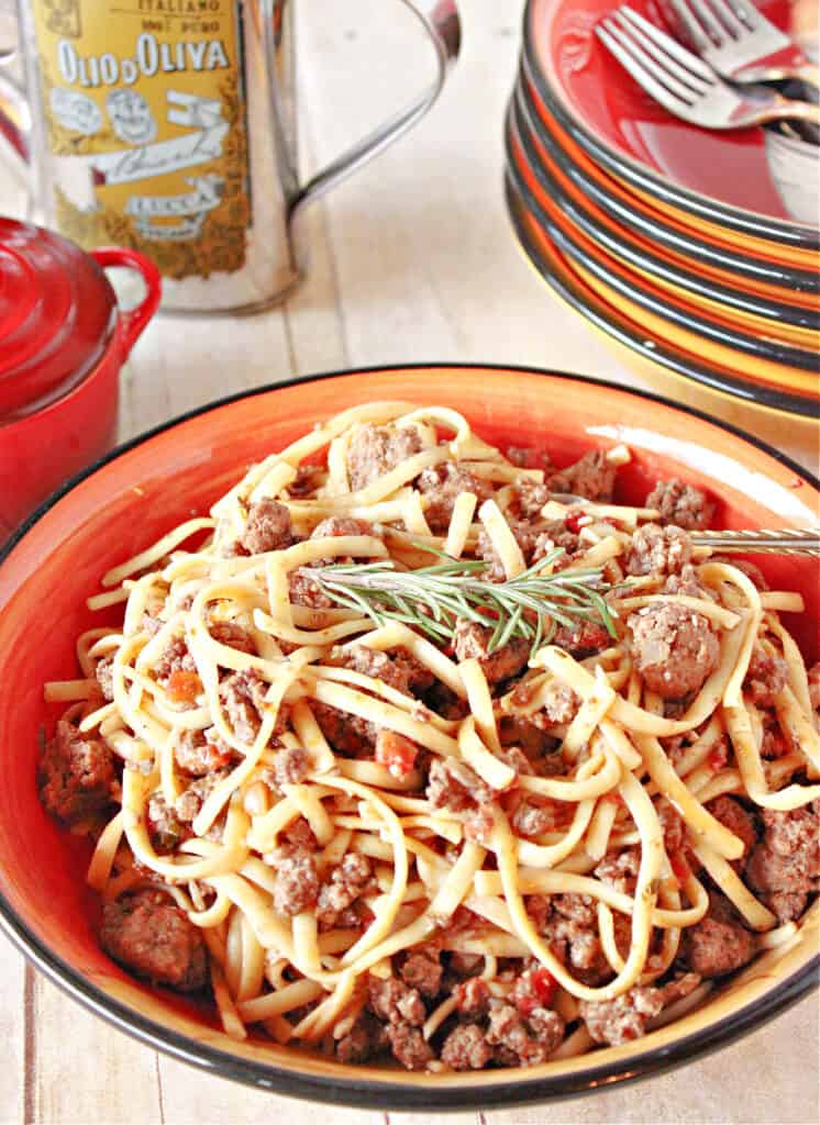 A vertical overhead closeup image of a bowl of Linguine with Lamb with a sprig of rosemary on top and a stack of bowls in the background.