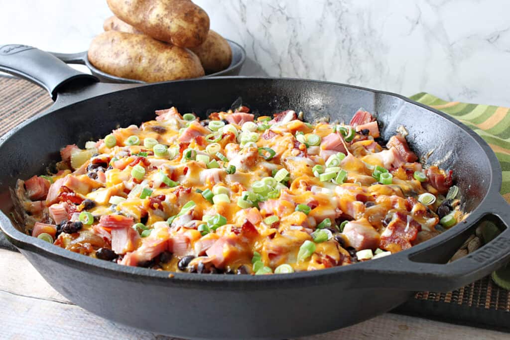 A horizontal photo of a black cast iron skillet filled with cheesy Loaded Skillet Potatoes topped with scallions along with some russet potatoes in the background.