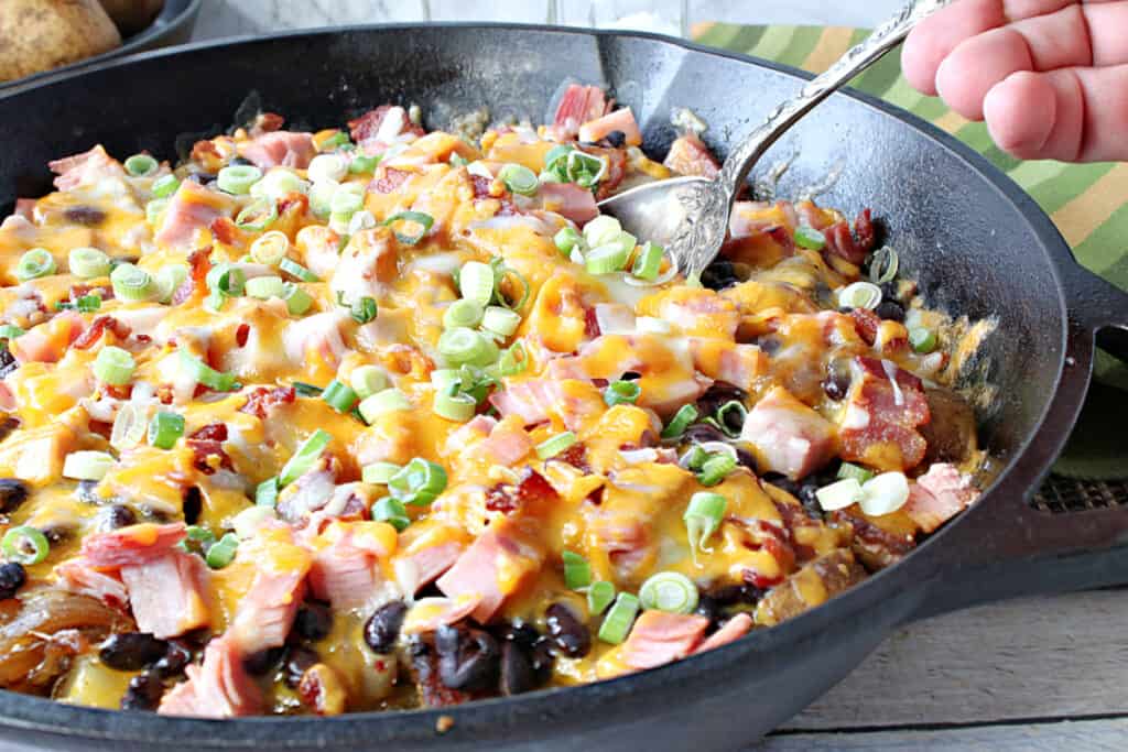 A horizontal photo of a cast iron skillet filled with Loaded Skillet Potatoes and a hand holding a serving spoon digging into the potatoes.