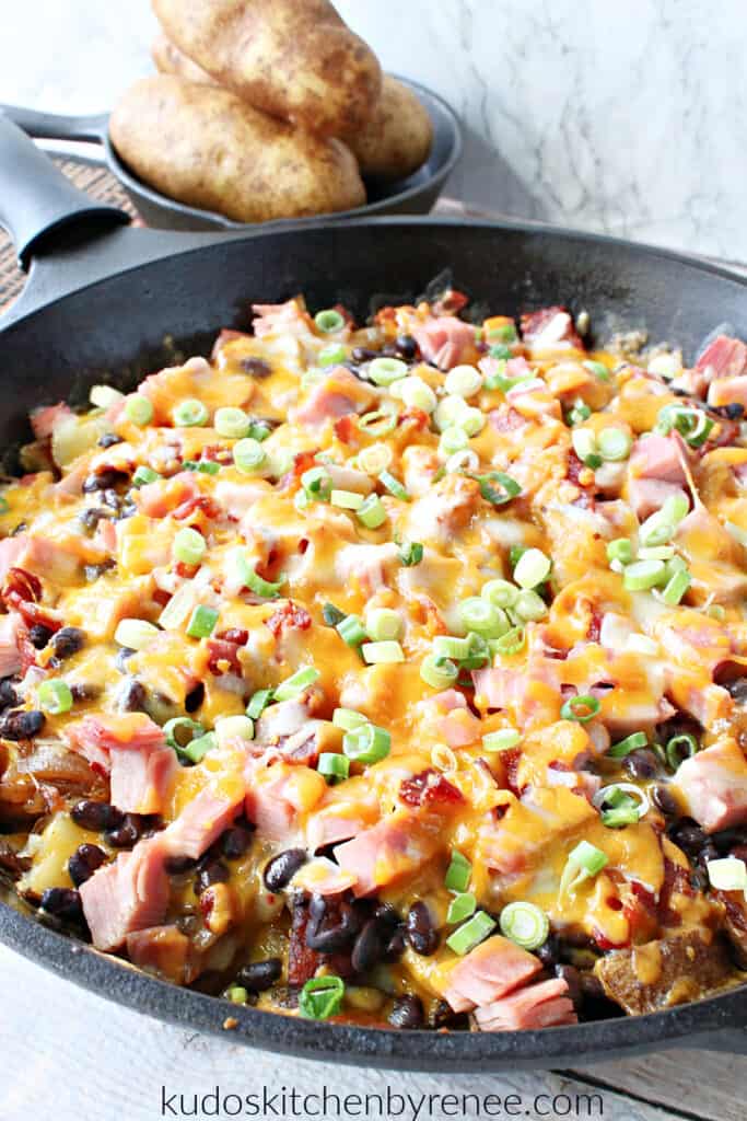 A vertical closeup image of a cast iron skillet filled with Loaded Skillet Potatoes with melted cheese, beans, ham, and scallions.