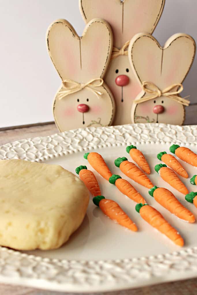 A vertical closeup image of a pretty platter with Homemade Marzipan Candy Carrots and a block of homemade marzipan along with some cute bunnies in the background.