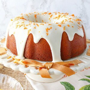 An Orange Bundt Cake with Bananas and Poppy Seeds on a white cake plate with an orange ribbon.