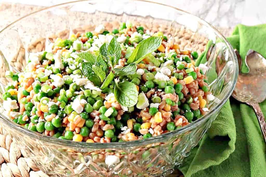 A horizontal offset photo of a glass bowl filled with Pea and Farro salad along with fresh mint and a green napkin on the side.