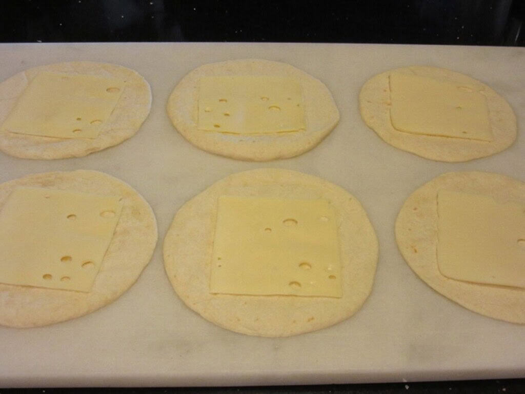Six flour quesadillas with six slices of Swiss cheese on a marble countertop.
