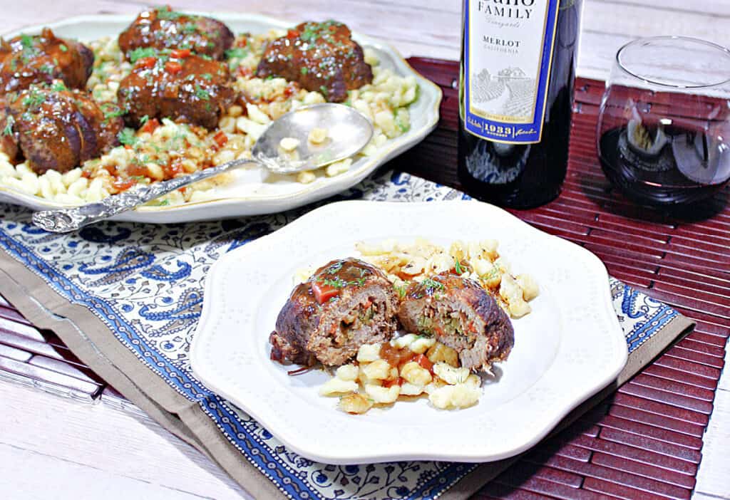 A dinner table set with a plate and a platter filled with Traditional German Beef Rouladen along with German spaetzle and a bottle of wine.