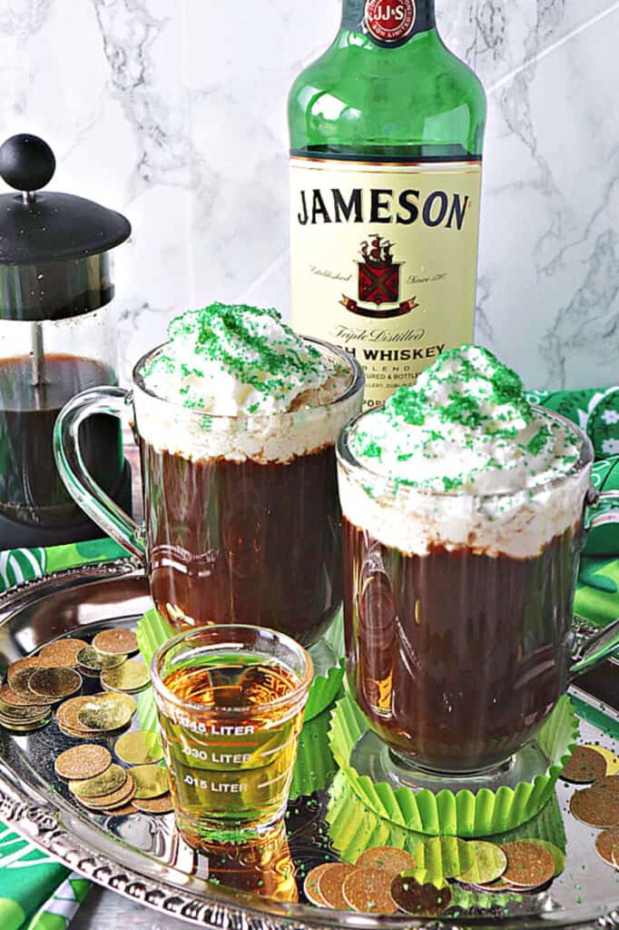 A vertical closeup of two mugs of Traditional Irish Coffee on a silver platter with gold coins and a shot of whiskey in the front.