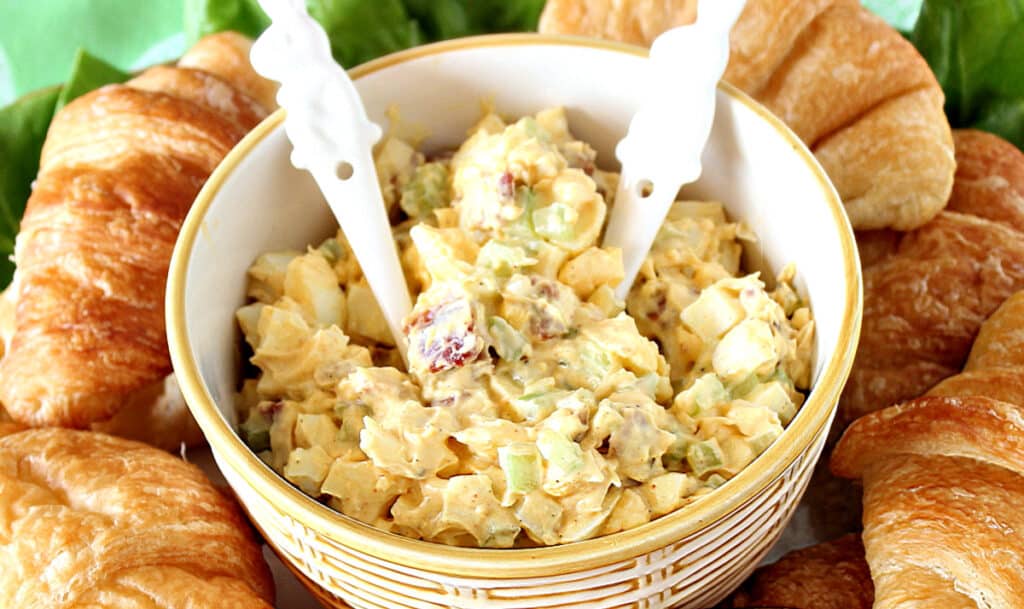 A overhead horizontal photo of Bacon Egg Salad in a yellow and white bowl with bunny spoons.