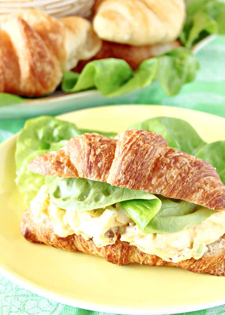A vertical closeup of Bacon Egg Salad on a croissant with butter lettuce on a yellow plate.