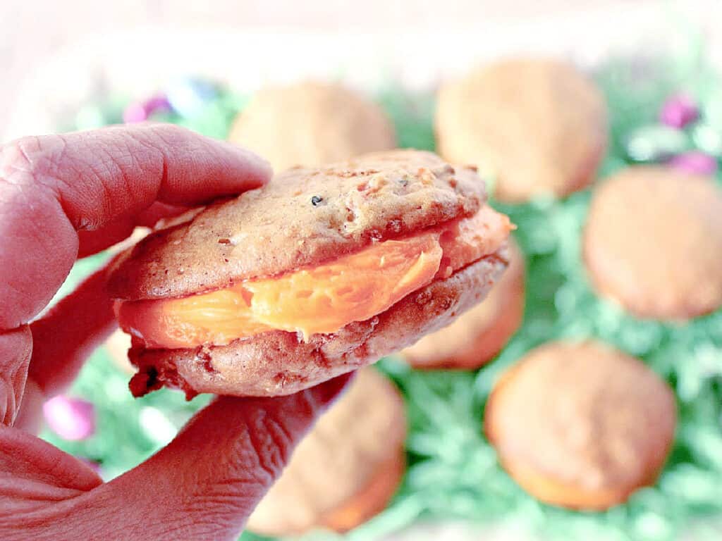 A closeup of a hand holding a Carrot Cake Whoopie Pie with orange cream cheese frosting in the middle.
