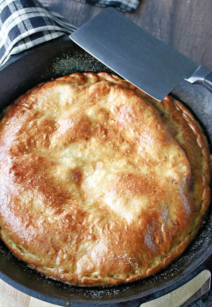 A vertical overhead closeup of a golden brown Double Crust Hawaiian Pizza in a cast iron skillet with a clever resting on the side.