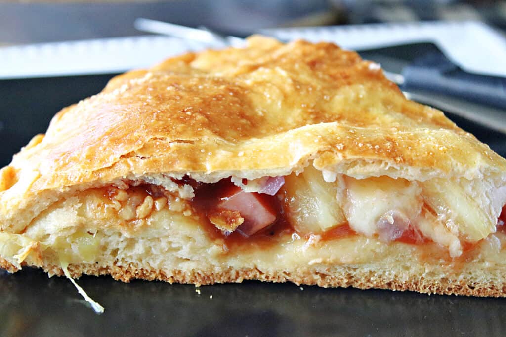 A super closeup of a slice of Double Crust Hawaiian Pizza with ham, pineapple, cheese, and red onion, on a black plate.