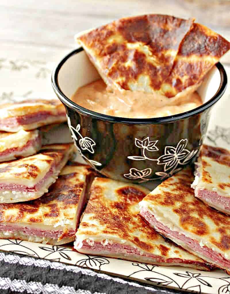 A vertical closeup of Reuben Quesadillas cut into triangles along with a center cup of 1000 Islands dipping sauce.