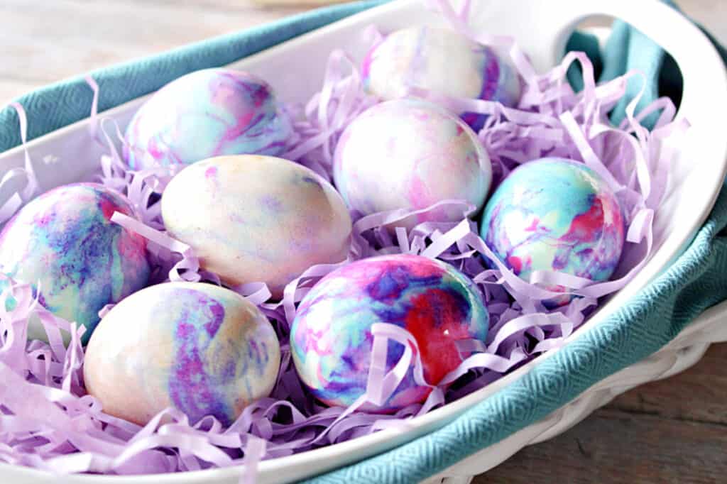 A horizontal photo of a white oval bowl of Tie-Dye Easter Eggs with a blue napkin underneath.