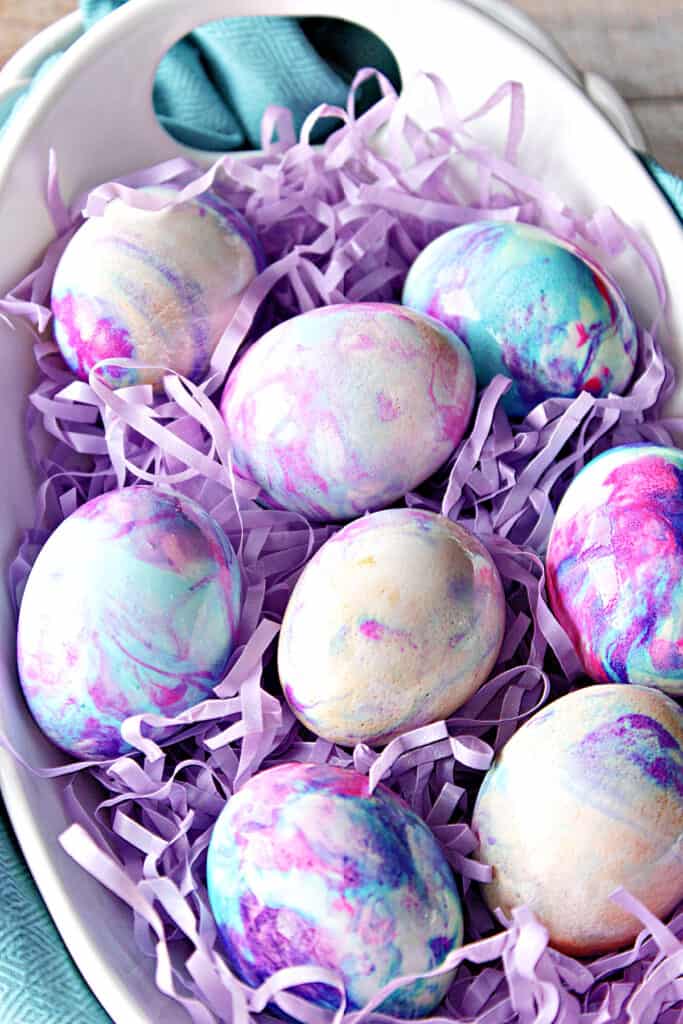 A vertical overhead closeup photo of pretty pastel Tie-Dye Easter Eggs in a white bowl with purple Easter grass.