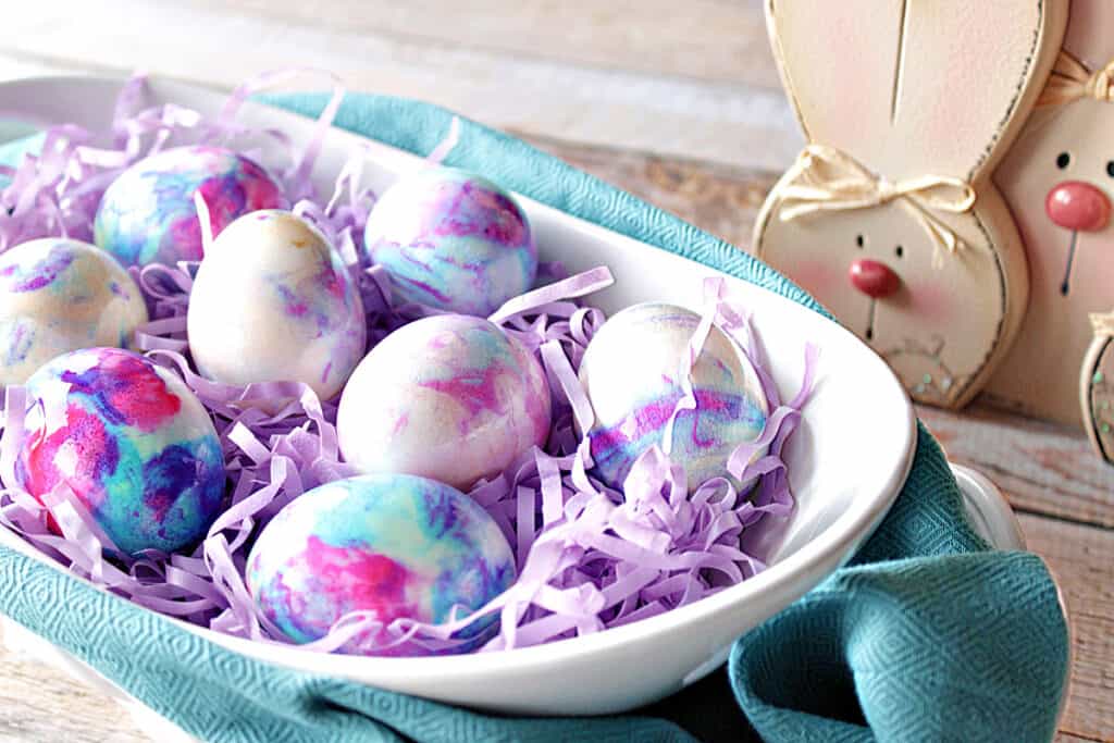 A white oval bowl with a blue napkin underneath filled with Tie-Dye Easter Eggs and cute wooden bunnies on the side.