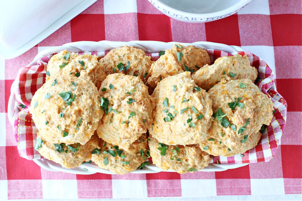 An overhead photo of a basket filled with a red and white checked napkin along with Cheddar Bay Biscuits topped with parsley.