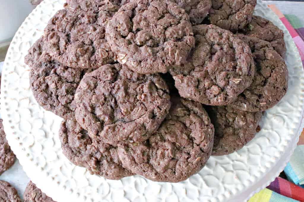 A closeup overhead photo of a pretty white cake plate filled with a pile of Chocolate Oatmeal Cookies.