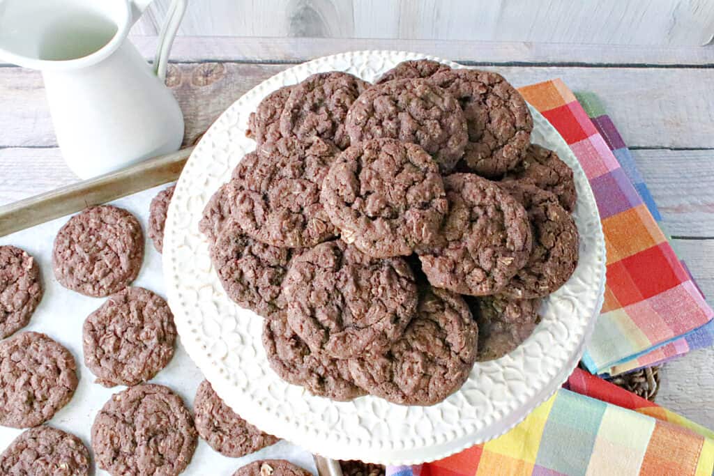 An overhead photo of a cake plate and a baking sheet filled with Chocolate Oatmeal Cookies along with a colorful napkin.